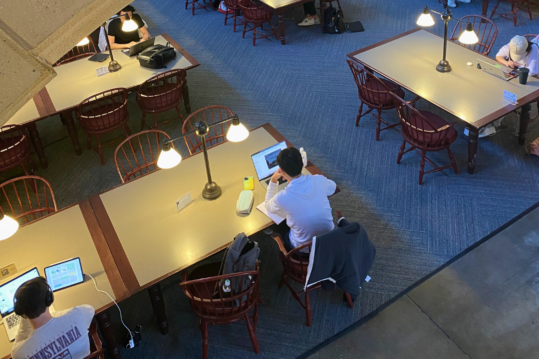 A cozy, well-lit study area with individuals at separate wooden tables, each equipped with a desk lamp. People are engaged in reading or working on laptops.
